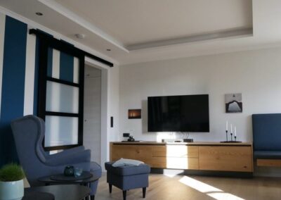 Wohnzimmer, Ohrensessel, Fernseher, Holzfußboden, schwarze Schiebetür, Bilder an der Wand, Sideboard unter dem Fernseher